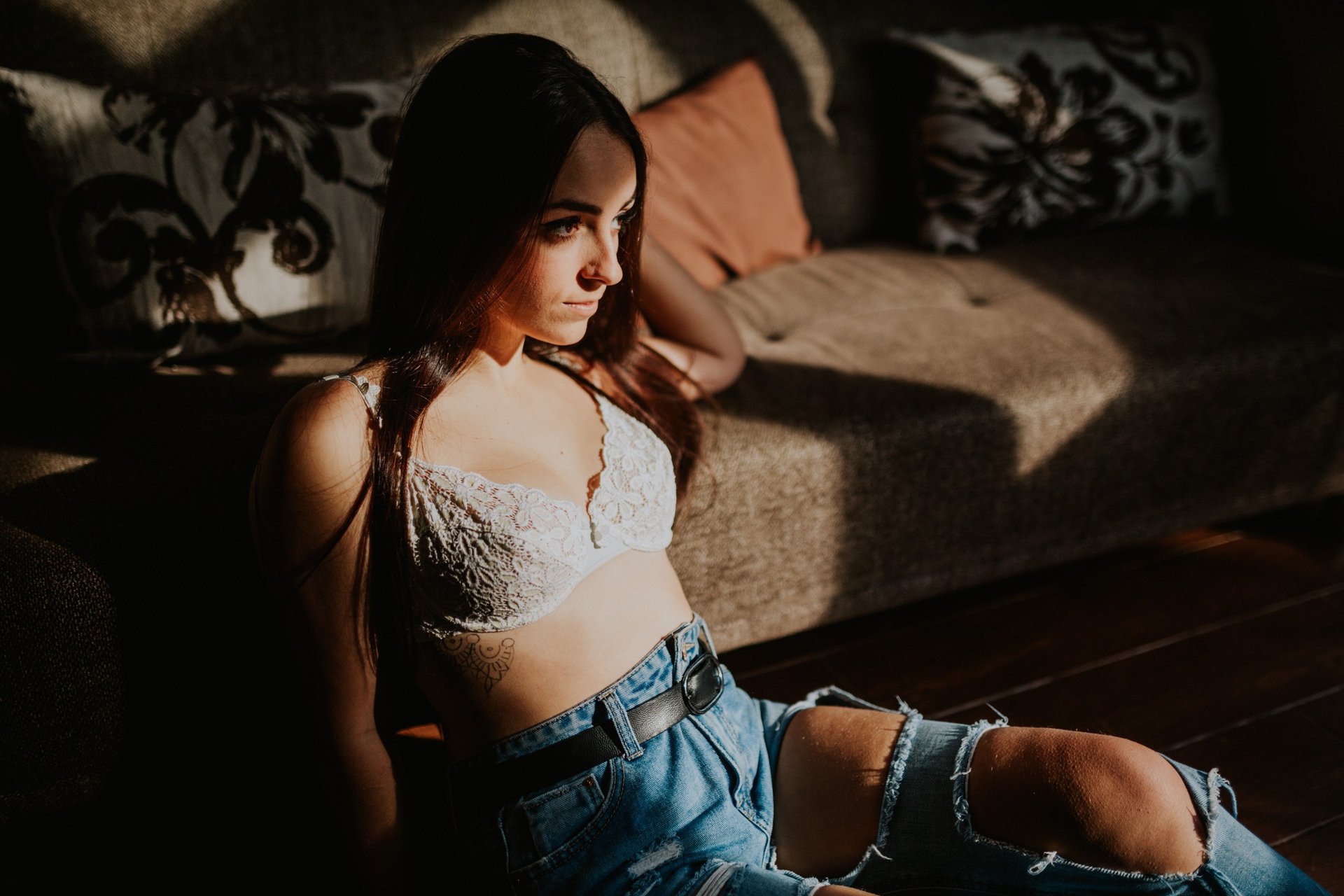 Mujer joven recostada en un sofá con luz natural lateral intensa, top de encaje blanco y shorts de mezclilla, actitud relajada y pensativa. Fotografía de boudoir de Víctor Ruiz, Granada.