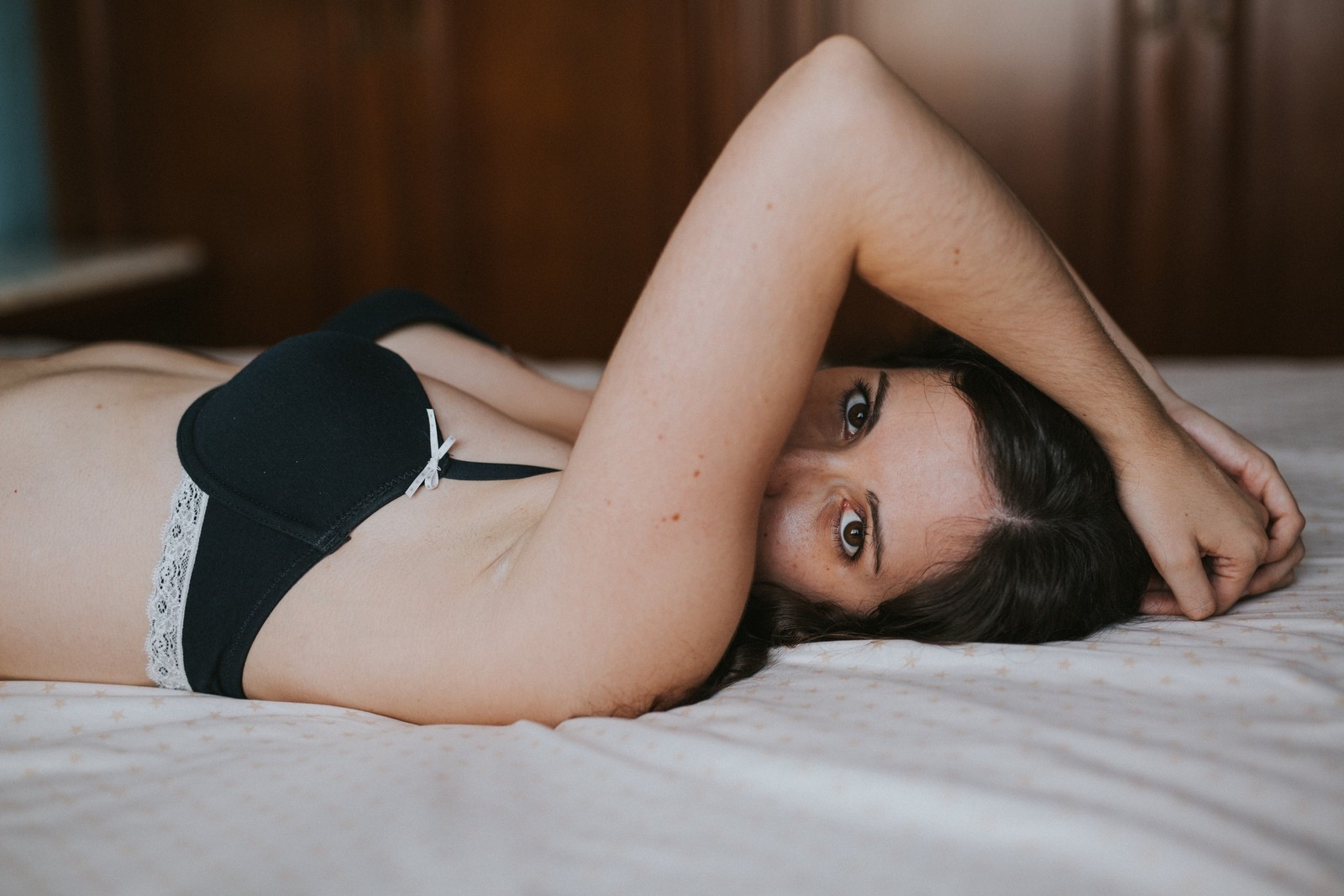 Mujer con cabello oscuro en lencería negra de encaje yaciendo en una cama de sábanas blancas, mirando a cámara con los brazos detrás de la cabeza. Luz natural suave, ambiente íntimo. Fotografía de boudoir de Víctor Ruiz, Granada.