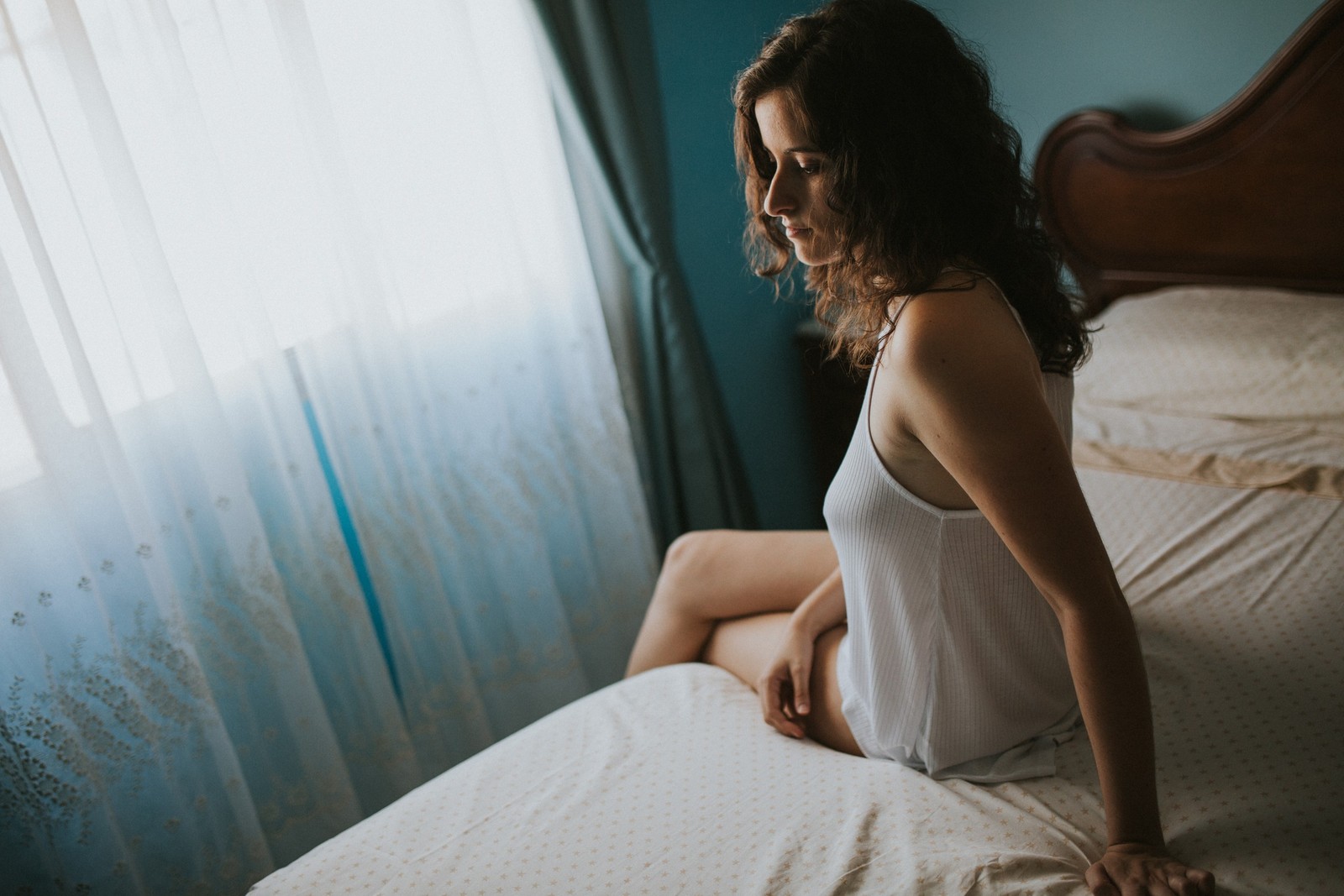 Mujer con camiseta blanca sin mangas sentada en el borde de una cama mirando hacia la ventana, con luz natural suave a través de visillos. Fotografía de boudoir de Víctor Ruiz, Granada.