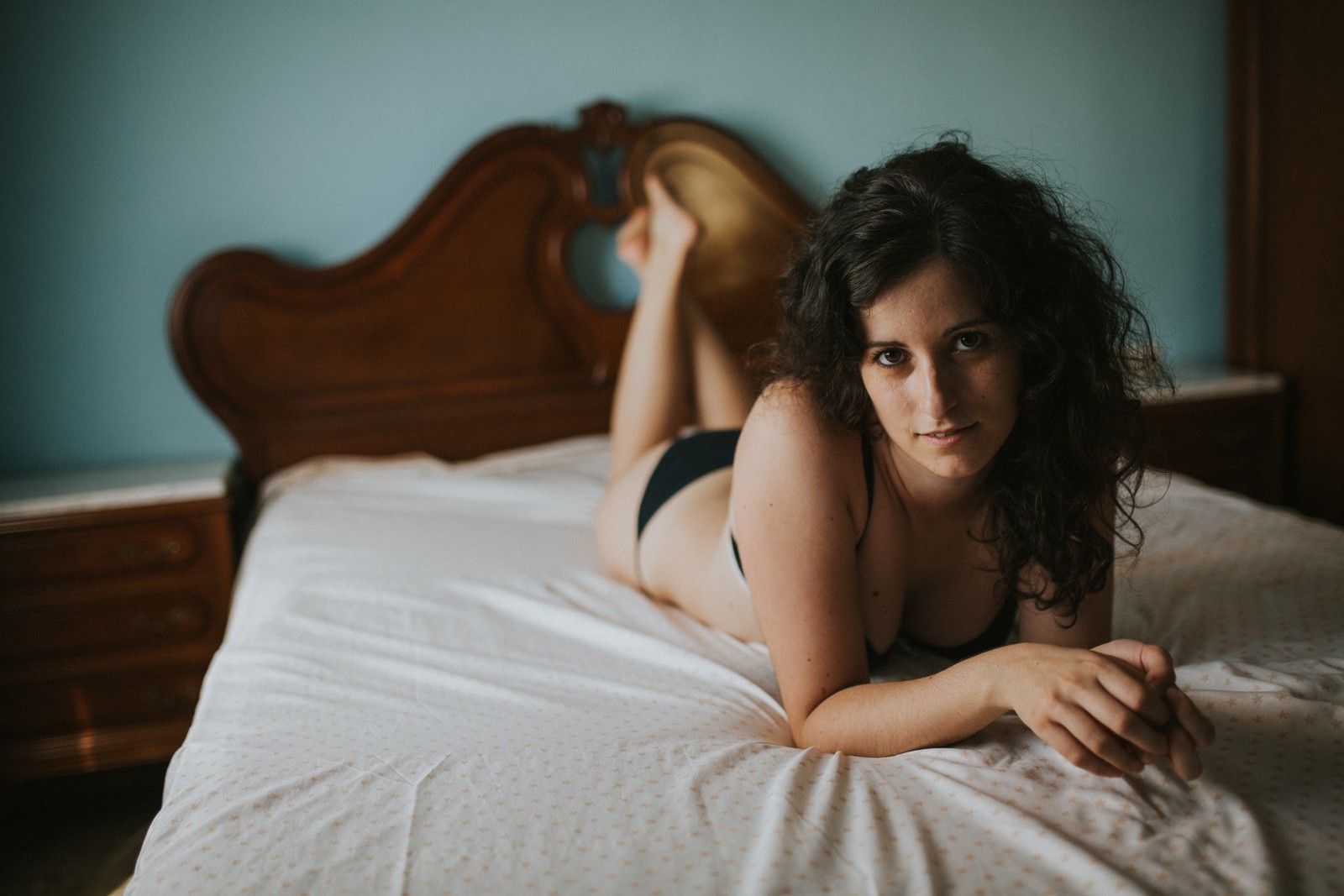 Mujer con cabello rizado yace boca abajo sobre una cama de matrimonio con lencería negra, apoyada en los codos mirando a cámara con expresión serena. Fotografía de boudoir de Víctor Ruiz, Granada.