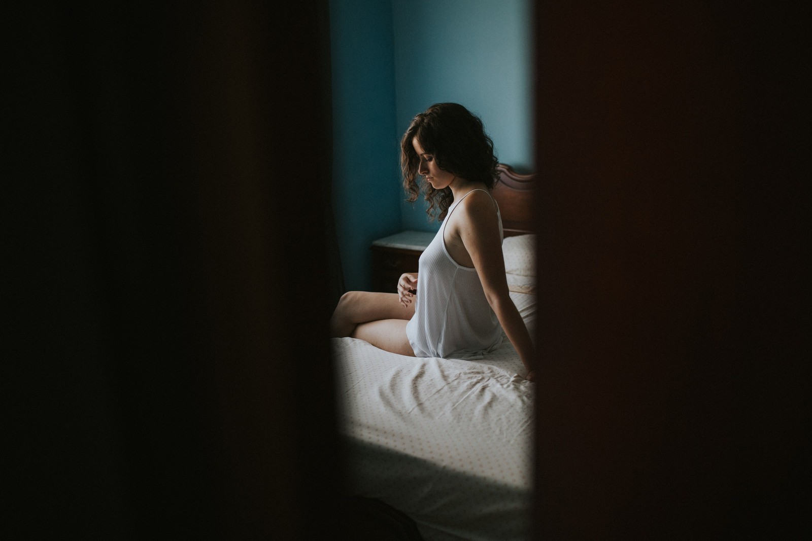 Mujer con camiseta blanca holgada sentada en la cama mirando hacia abajo en actitud reflexiva, en una habitación con paredes azules y luz natural tenue. Fotografía de boudoir de Víctor Ruiz, Granada.