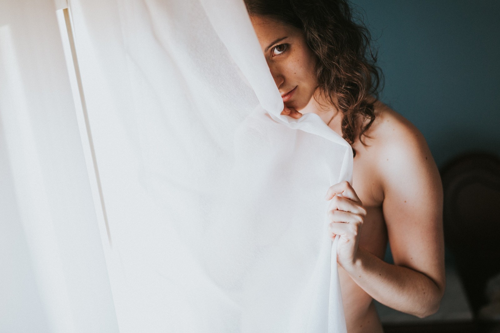 Mujer con cabello rizado sosteniendo una cortina blanca translúcida frente al rostro, mirando a cámara, iluminada por luz de ventana. Fotografía de boudoir de Víctor Ruiz, Granada.