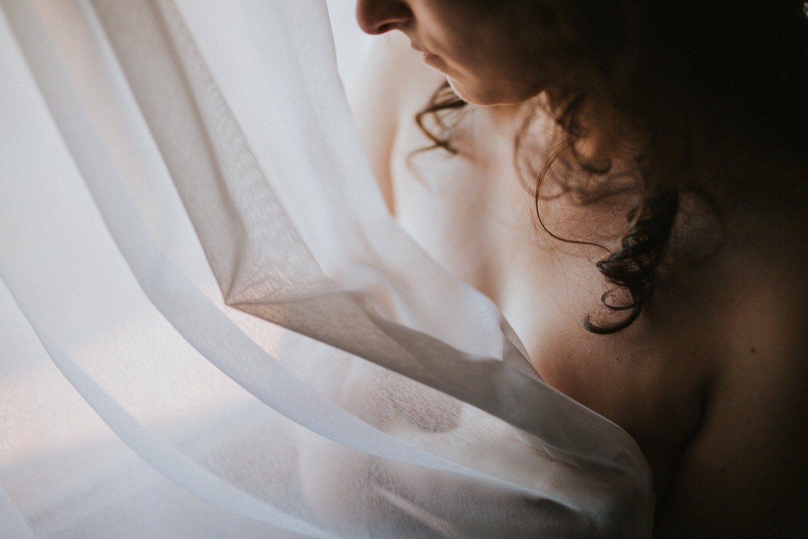 Primer plano de una mujer de perfil cubierta parcialmente por una tela blanca vaporosa, luz natural suave que la hace brillar. Fotografía de boudoir de Víctor Ruiz, Granada.
