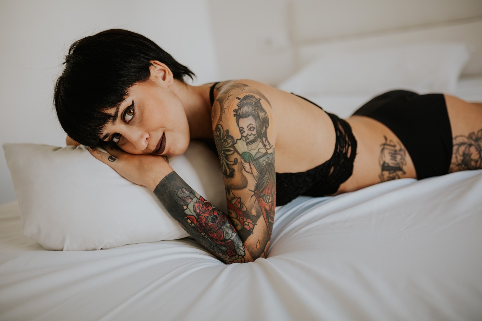 Mujer con cabello corto oscuro y tatuajes recostada boca abajo sobre una cama blanca con lencería negra de encaje, mirando a cámara, luz natural suave. Fotografía de boudoir de Víctor Ruiz, Granada.