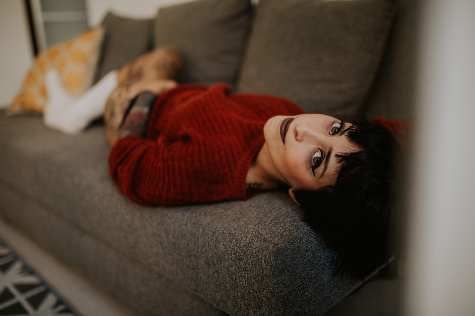 Mujer con cabello corto y tatuajes recostada en un sofá gris con un suéter rojo y calcetines blancos, ambiente acogedor e íntimo. Fotografía de boudoir de Víctor Ruiz, Granada.